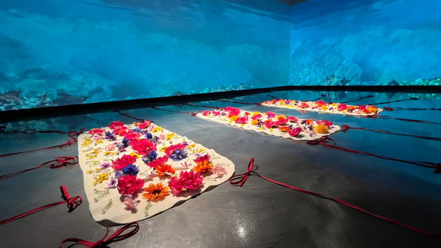 A projection of the floor of the Aegean Sea makes the walls blue and on the floor lies three mats with brightly colored flowers placed on them