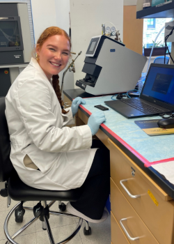 A woman wearing gloves and a lab coat smiles next to her workstation at the Singh laboratory.