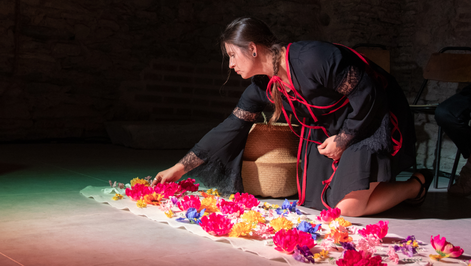 A woman kneels on the floor in a well-lit performance space and places flowers on the ground in a pattern as part of an art performance.