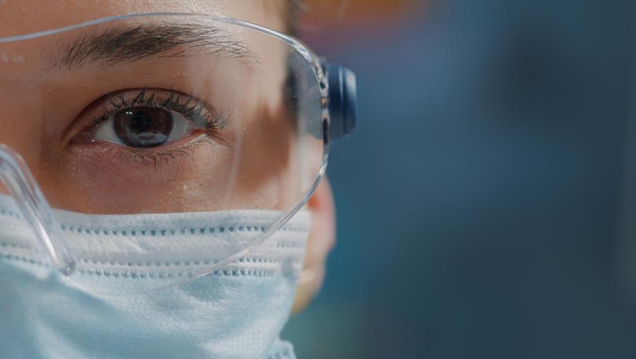 Scientist showing one eye on camera in laboratory, wearing safety glasses and face mask. Woman biologist looking at camera with face of face, working in research lab. 