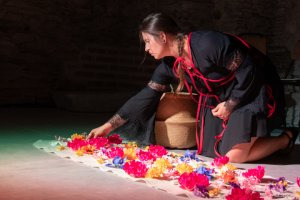 A woman kneels on the ground in a well lit performance art space. She is placing brightly colored flowers on the ground in a pattern.